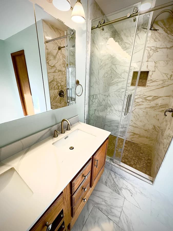 Oak vanity with a white quartz-look top facing the sliding glass shower over marble-look floor tile.