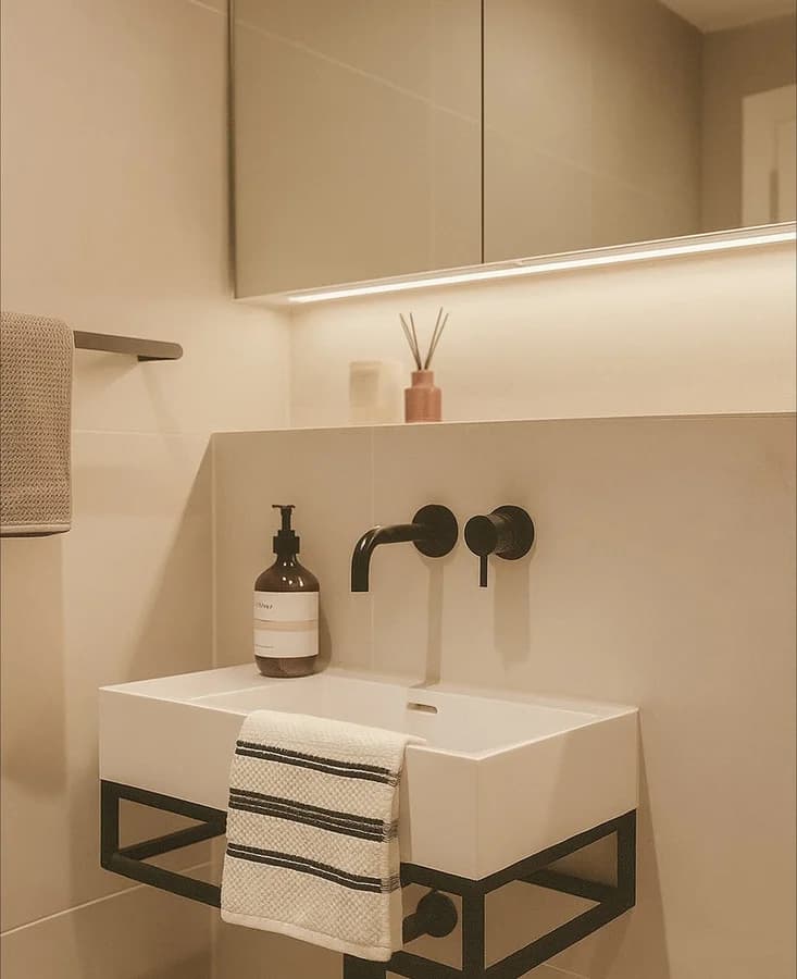 Wall-mounted black mixer over a rectangular basin on a black frame, below a lit mirror cabinet.