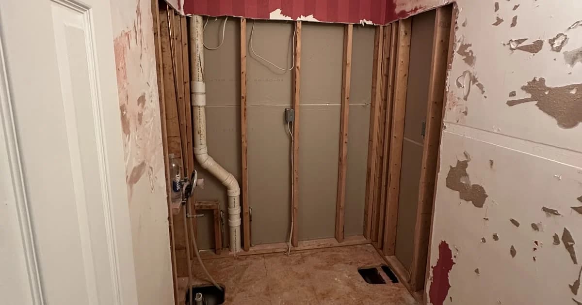 A bathroom opened to the studs during demolition: wall framing, the drain riser and a cleared subfloor.