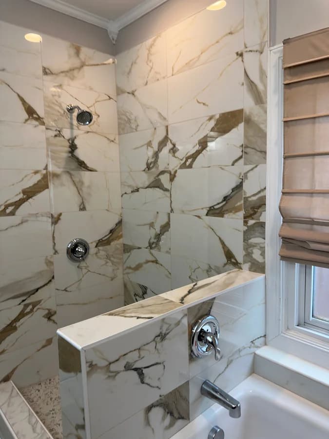 Shower wall in large-format marble-look tile with a chrome shower head and valve above a tiled pony wall.