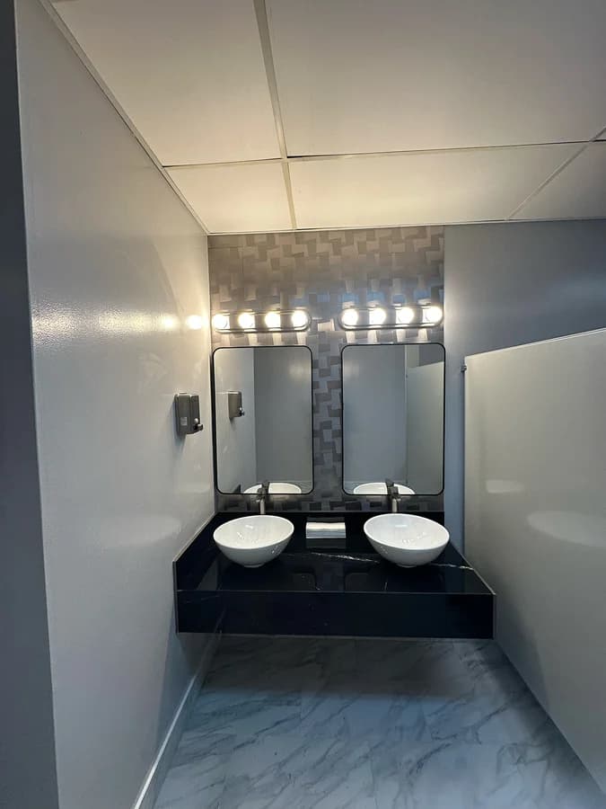 Restroom seen from the entrance: twin vessel basins, privacy partition and marble-look floor tile.