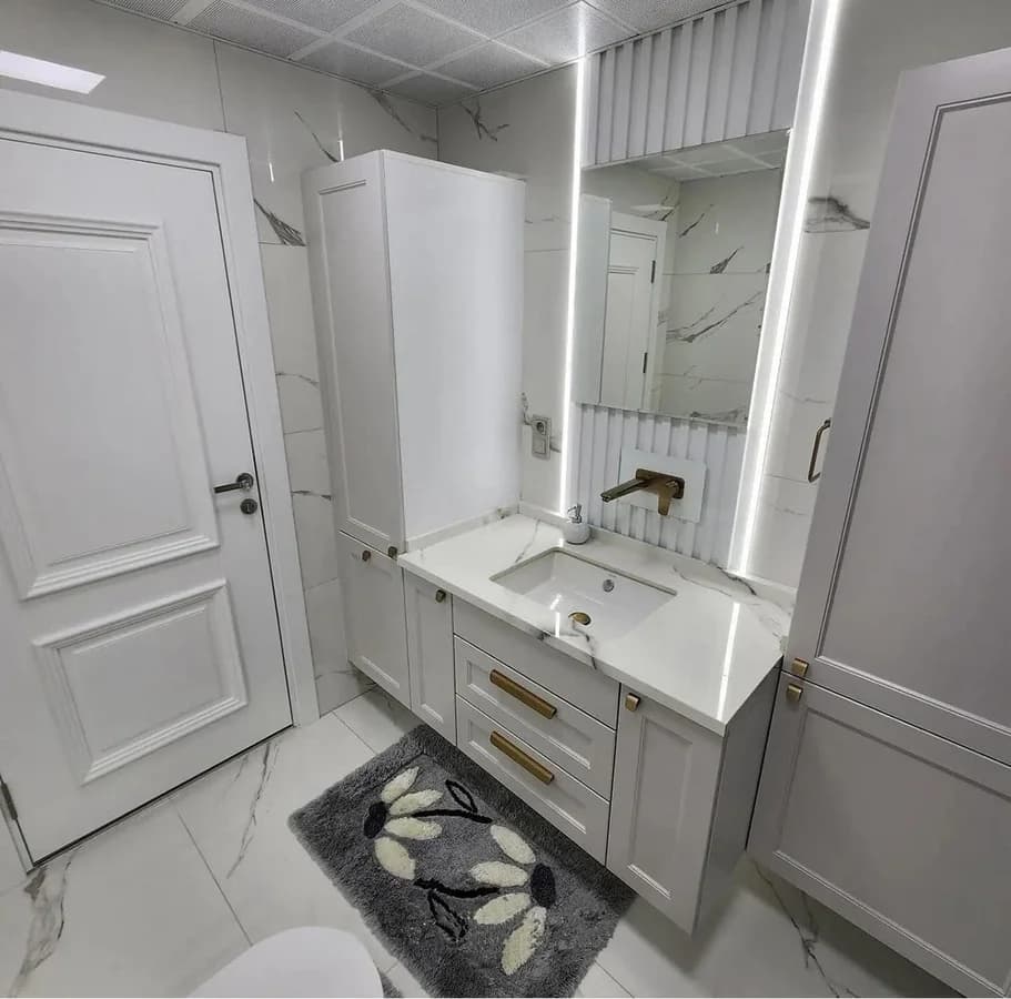 Vanity under a lit mirror with a brass wall-mounted mixer and a fluted backsplash panel.