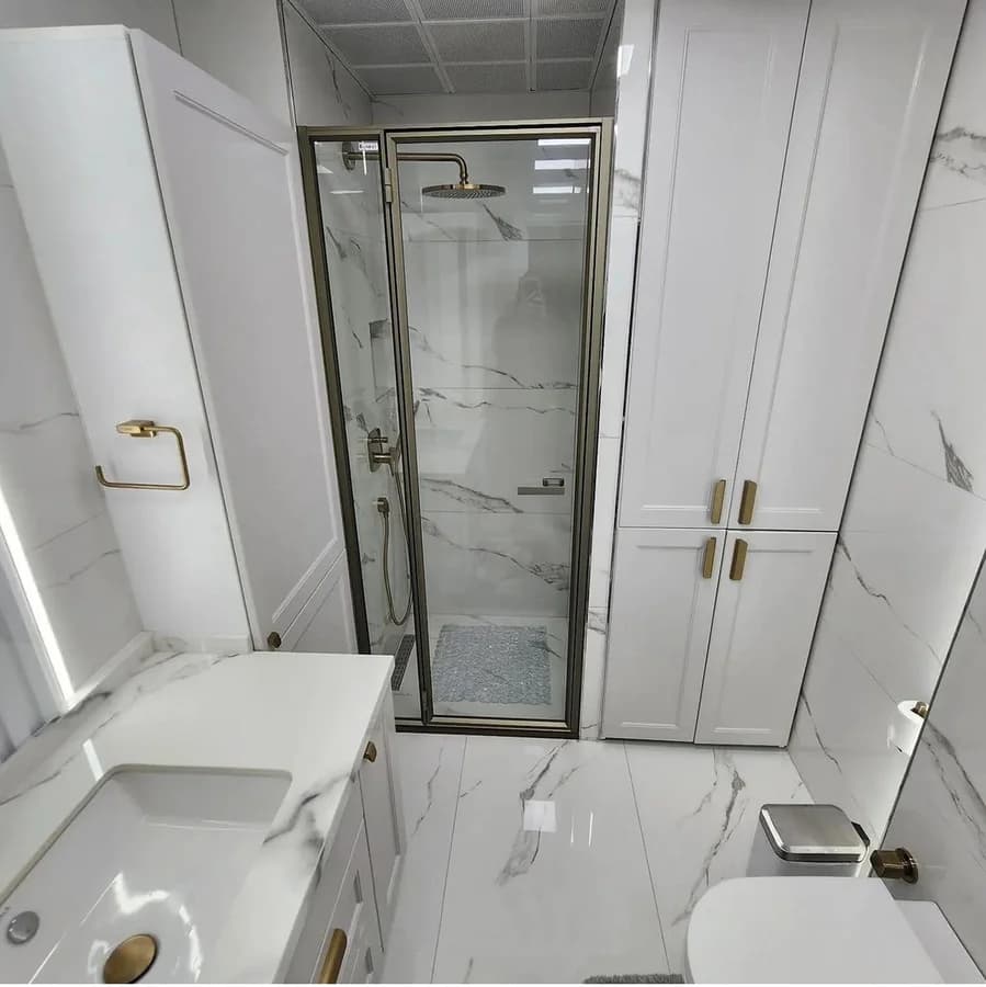 Framed glass shower with a brass rain head and pebble floor, between tall white cabinets.
